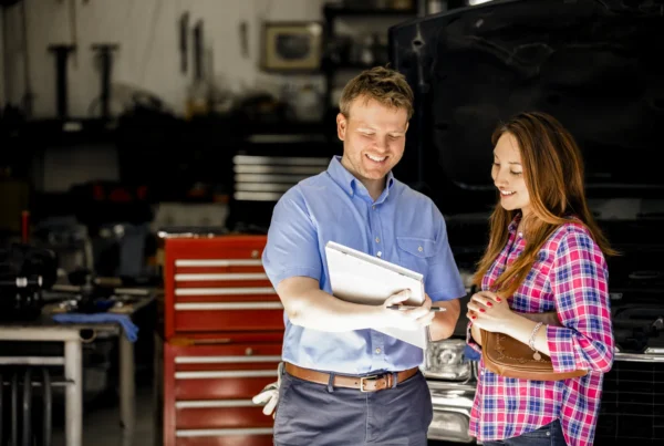 A smiling auto shop service advisor holding a clipboard reviews it with a female customer inside a repair bay with a red tool chest and a car with its hood open.