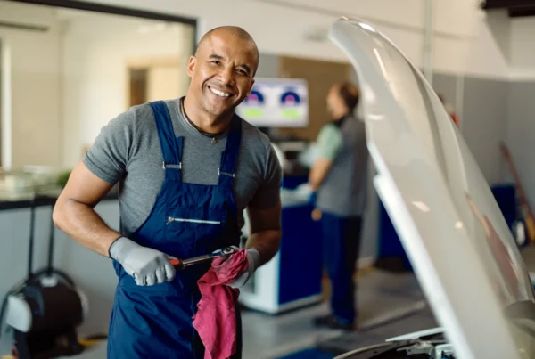 A smiling mechanic in navy overalls holds a wrench and red rag next to a car with its hood open inside an auto repair shop.