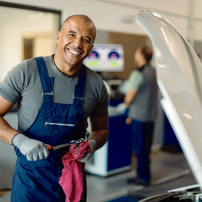 A smiling mechanic in navy overalls holds a wrench and red rag next to a car with its hood open inside an auto repair shop.